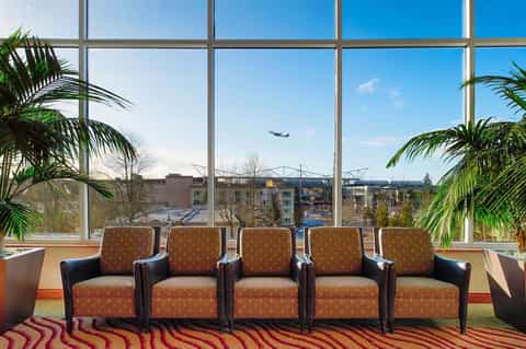 Modern lounge with five orange patterned chairs facing floor-to-ceiling windows overlooking cityscape and airplane