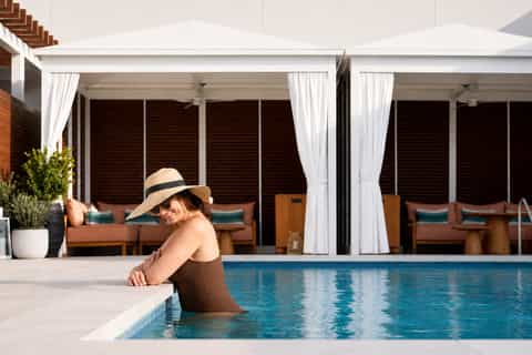 Woman in hat swimming in indoor pool with white cabanas and lounge chairs with brown cushions visible in background