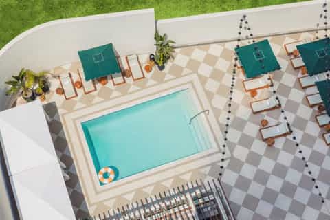 Aerial view of resort pool surrounded by teal umbrellas, wooden lounge chairs, decorative pavers, and white walls