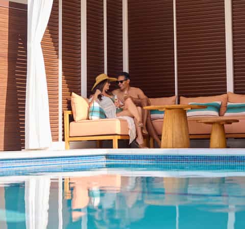 Couple relaxing on peach cushioned lounge chair with wooden arms by resort pool, holding drinks under brown louvered shade