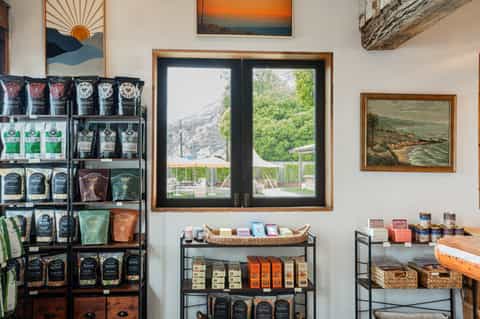 Retail shop interior with wooden-framed windows, mountain view, tiered shelving with packaged products, and landscape artwork