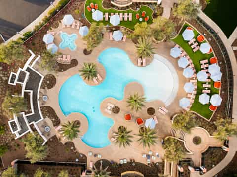 Aerial view of resort pool complex with curved pools, pink lounge chairs, umbrellas, and surrounding palm trees
