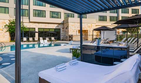 Contemporary pool deck with white lounge chairs, black pergola shade structures, and hotel building