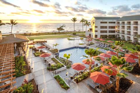Beachfront resort at sunset with pool, red umbrellas, palm trees, and ocean view