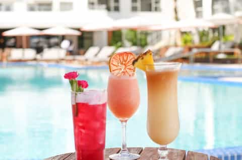 Three colorful cocktails by poolside: red drink with flower, pink grapefruit cocktail, and tropical drink with pineapple