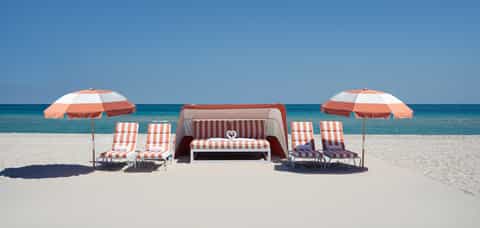 Beach setup with red and white umbrellas, striped chairs, lounge bed, and ocean view