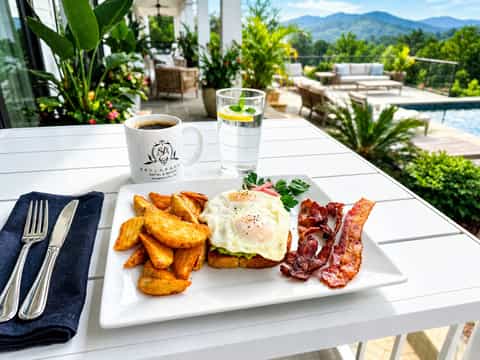 Breakfast plate with fried eggs, bacon, and potatoes on a terrace overlooking mountains and pool