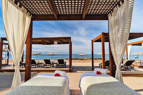 Beachfront massage beds with white linens under wooden pergolas overlooking ocean and distant mountains