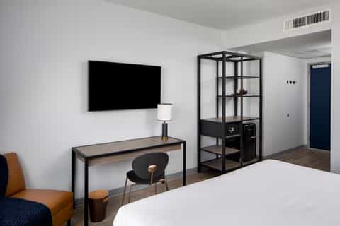 Modern hotel room with white walls, dark desk with TV, black industrial shelving unit, tan accent chair, and minimalist decor