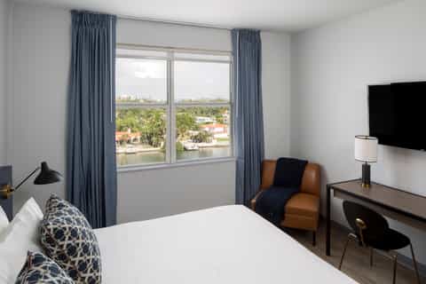 Bright hotel room with large windows overlooking water, blue curtains, white bed with patterned pillows, and tan accent chair