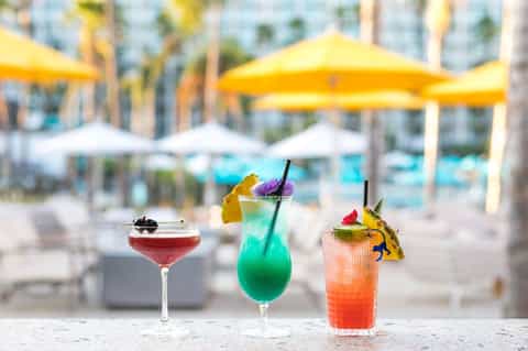 Three colorful cocktails with fruit garnishes poolside, yellow umbrellas blurred in background