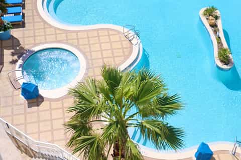 Aerial view of resort pools with palm tree island feature and blue decking surrounded by lounge chairs