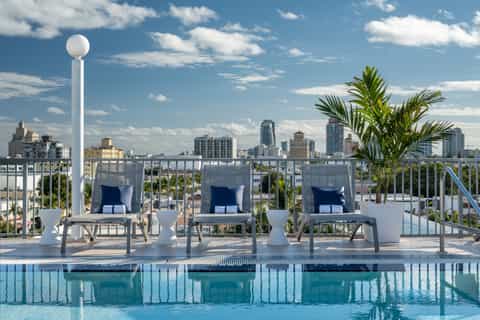 Rooftop pool deck with loungers, tropical plants, and panoramic city skyline views on clear blue day