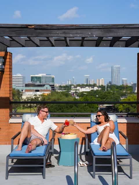 Couple toasting colorful cocktails on rooftop terrace with city skyline view and pergola overhead