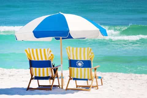 Two yellow and white striped beach chairs under blue and white umbrella facing turquoise ocean waves