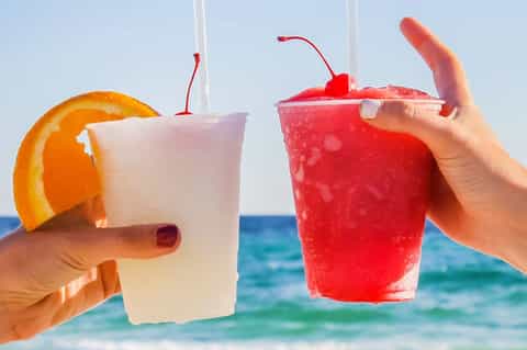 Two tropical cocktails with orange slice and cherry garnish held against ocean backdrop