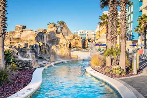 Resort lazy river with cascading waterfall and rock formations surrounded by palm trees and colorful buildings