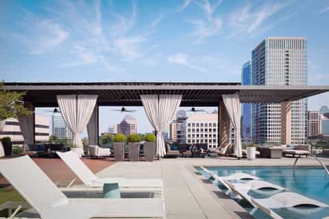 Rooftop lounge with pergola, curtains, lounge seating, infinity pool, and downtown skyline at daytime