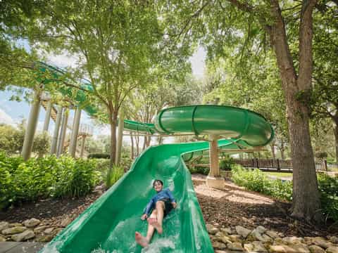 Child sliding down green water slide in wooded park setting with trees and playground equipment