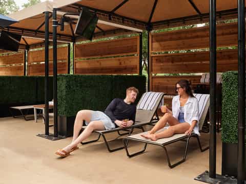 Two guests relaxing in lounge chairs under pergola with tan canopy and green artificial wall panels