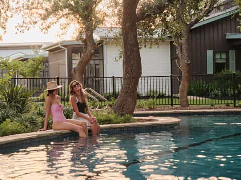 Two women sitting poolside under large oak tree with resort cabins visible in background