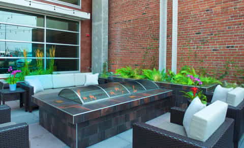 Modern outdoor patio with dark fire pit, white cushioned lounge chairs, and brick building backdrop with purple flowers