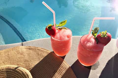 Two pink cocktails with strawberries and straws poolside with ocean view in background