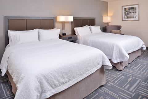 Two queen beds with white bedding in a modern hotel room with brown headboards and patterned carpet