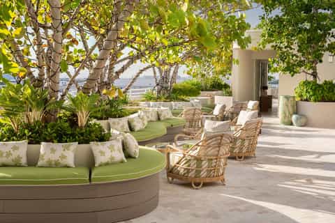 Shaded rooftop terrace with curved green seating, rattan chairs, tropical plants, and ocean views