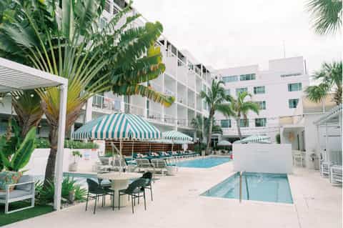 Resort pool area with striped umbrellas, white lounge chairs, and multi-story hotel building backdrop