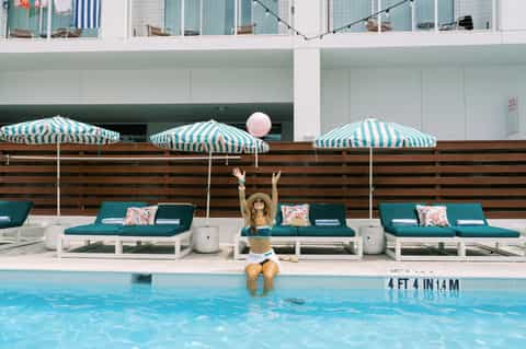 Woman sitting poolside under teal striped umbrellas with lounge chairs and modern hotel building