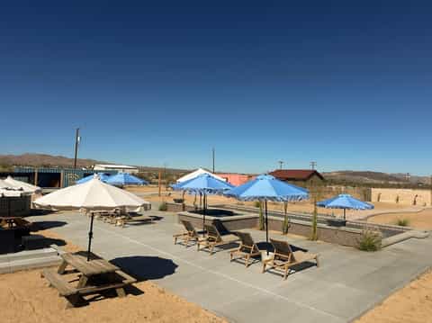 Resort pool deck with blue and white umbrellas, lounge chairs, and desert mountain views