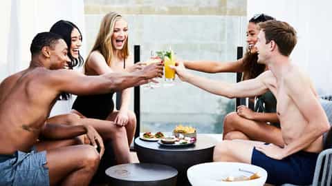 Group of six young adults toasting with colorful drinks at a rooftop lounge with city views and appetizers