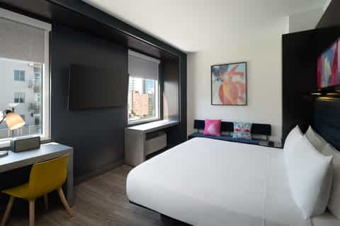 Hotel bedroom with queen bed, black accent wall, yellow desk chair, and city skyline views