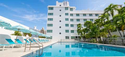 Resort pool with bright turquoise water, white loungers, umbrellas, palm trees, and Art Deco-style hotel tower