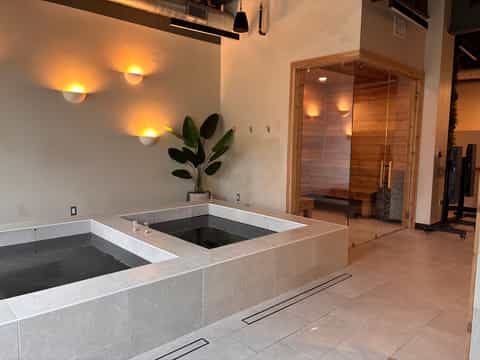 Spa relaxation area with two soaking tubs, wooden sauna with glass doors, warm accent lighting, and potted plant