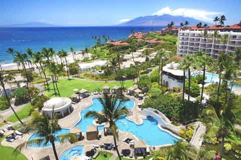 Aerial view of beachfront resort with curved pools, palm trees, and West Maui mountain in background