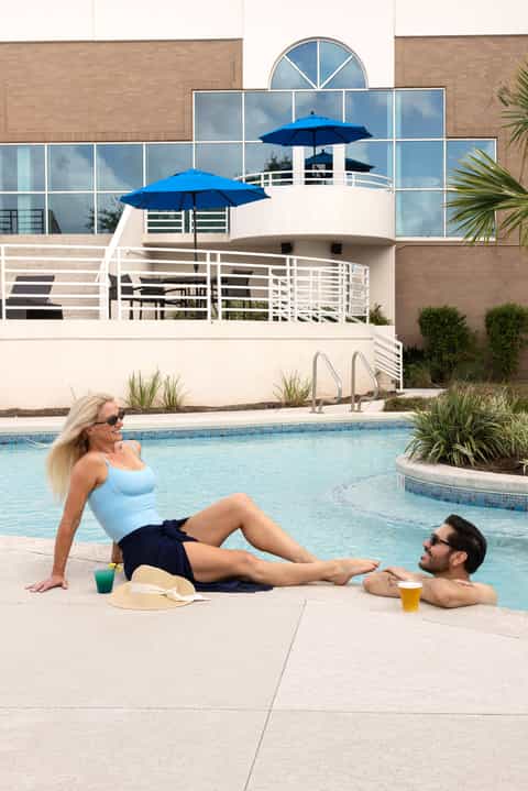 Woman and man relaxing poolside in front of a modern white building with blue umbrellas and glass balcony doors