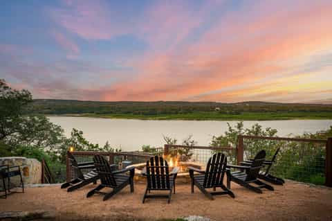 Sunset over a tranquil lake with Adirondack chairs arranged around an outdoor fire pit