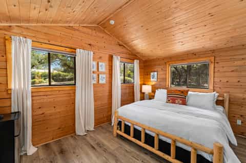 Cozy wooden cabin bedroom with queen bed, white bedding, and multiple windows with views