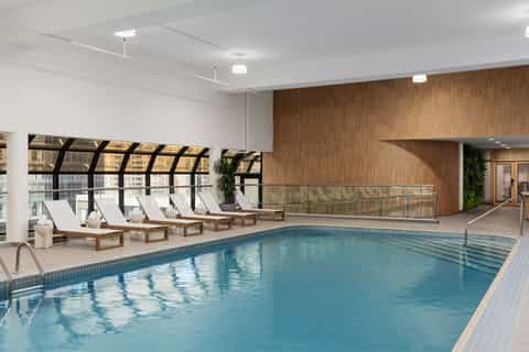 Indoor pool with wooden loungers, white cushions, and floor-to-ceiling windows in luxury facility