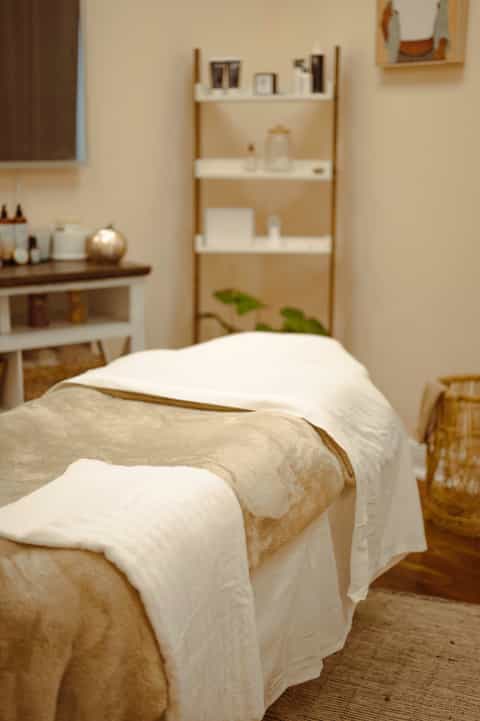 Minimalist spa treatment room with beige walls, wooden shelving, vanity, and massage bed
