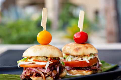 Two gourmet sliders with pulled meat, topped with colorful cherry tomatoes on wooden skewers, served on a dark plate
