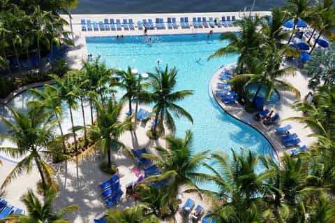 Aerial view of large resort pool with blue loungers, palm trees, and ocean backdrop