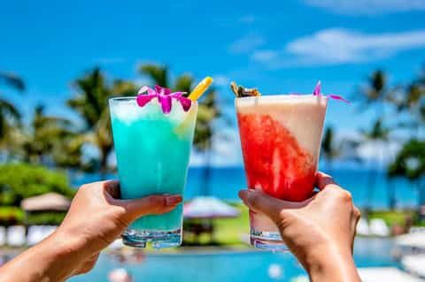 Two colorful tropical cocktails with flowers and straws held over a pool and palm trees
