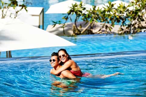 Couple in sunglasses enjoying crystalline blue swimming pool with white umbrellas and tropical landscaping