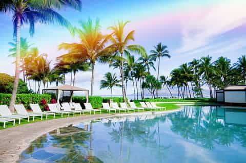 Tropical resort pool with palm trees, white lounge chairs, and ocean view at sunset