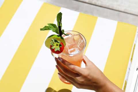 Hand holding tropical cocktail with kiwi and mint garnish against yellow striped background