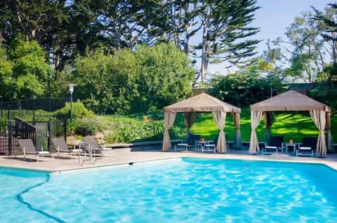 Resort outdoor pool with crystal blue water, lounge chairs, and beige gazebos surrounded by green landscaping and tall cypress trees