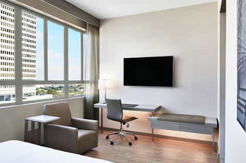 Modern hotel room with desk, office chair, flat-screen TV, armchair, and floor-to-ceiling windows overlooking city